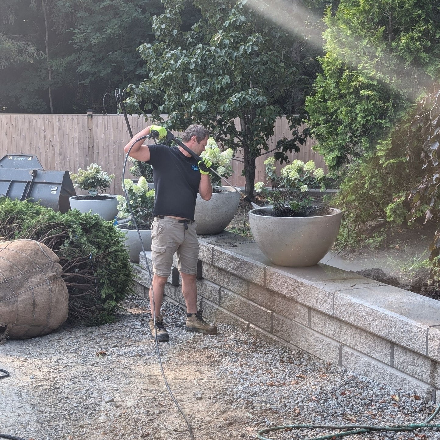 Person using a pressure washer in a garden setting with plants and a stone wall.
