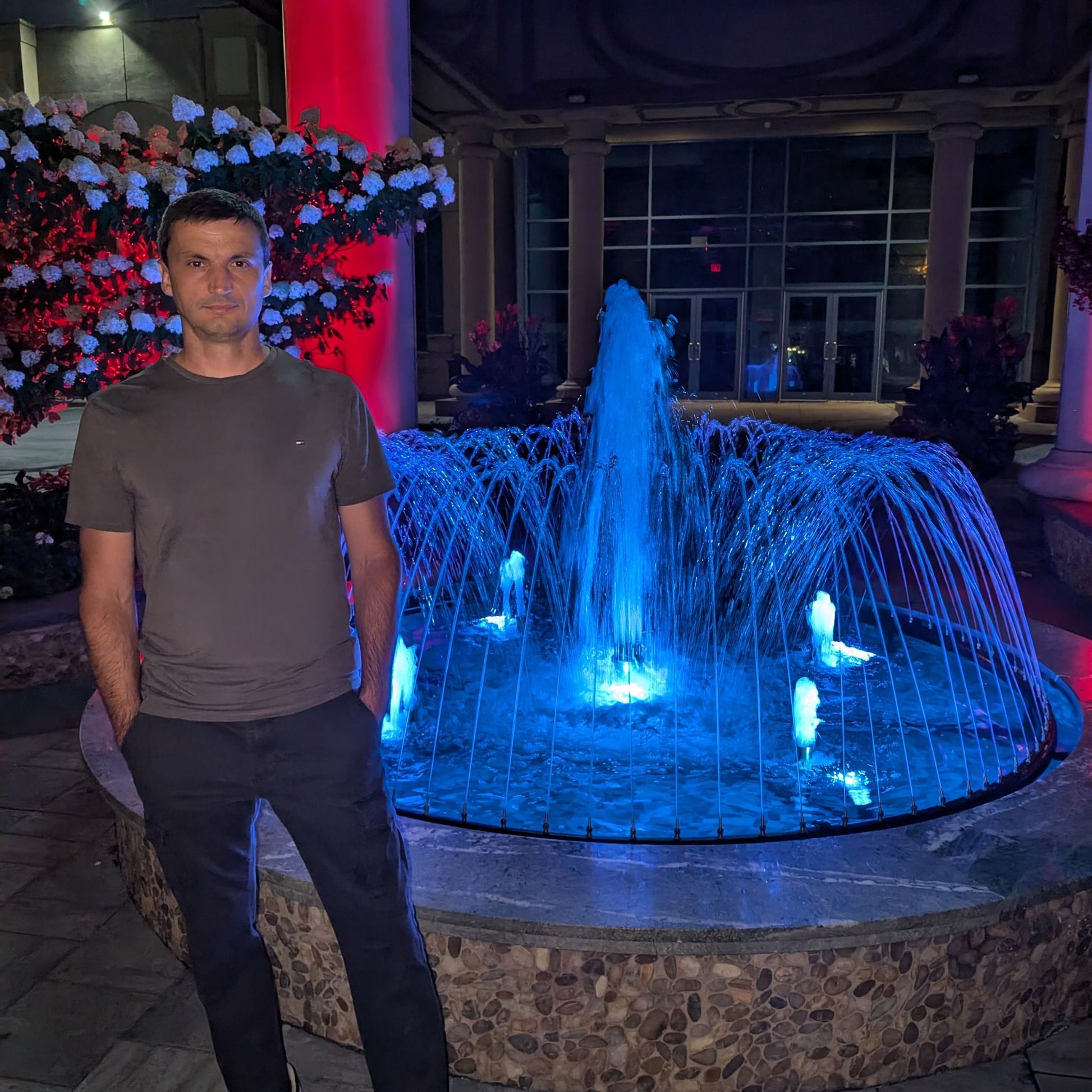 Man standing in front of a blue illuminated fountain at night.