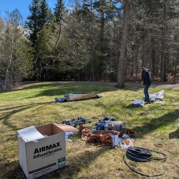 An outdoor scene in a wooded clearing shows a large white AIRMAX Diffused Aeration box in the foreground, with equipment and parts scattered on the grass, a tent laid down in the distance, and a person standing near trees on the right.