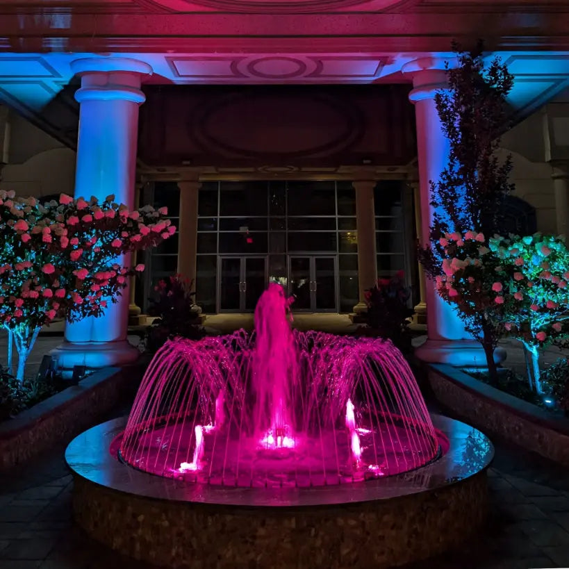 Fountain illuminated with pink light in front of a building with columns and floral decorations.