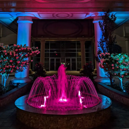 Fountain illuminated with pink light in front of a building with columns and floral decorations.
