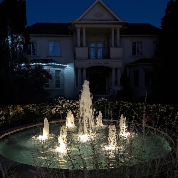 An illuminated outdoor circular fountain at night in front of a large house, with water jets lit from below and dark landscaping around the pool.