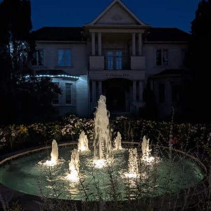 An illuminated outdoor circular fountain at night in front of a large house, with water jets lit from below and dark landscaping around the pool.