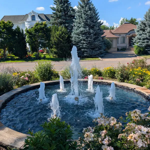 A circular outdoor fountain with multiple water jets in a landscaped garden bed, surrounded by greenery, flowers, and a suburban street with houses in the background.