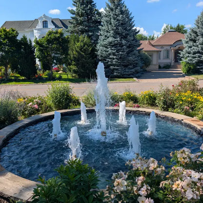 A circular outdoor fountain with multiple water jets in a landscaped garden bed, surrounded by greenery, flowers, and a suburban street with houses in the background.