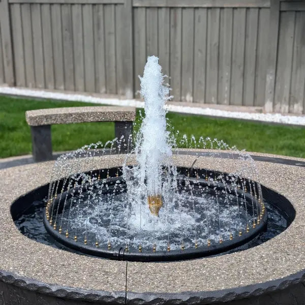 A round outdoor fountain with a central high jet and surrounding spray, set in a stone basin with a wooden fence and grass in the background.