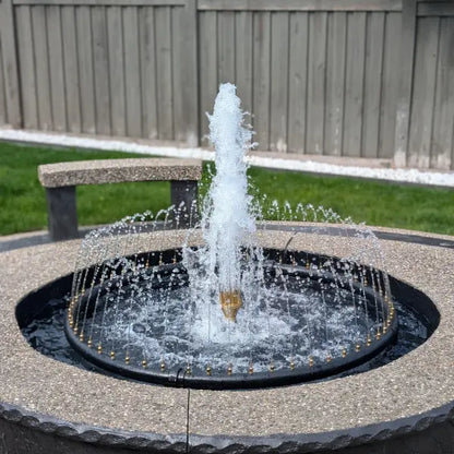 A round outdoor fountain with a central high jet and surrounding spray, set in a stone basin with a wooden fence and grass in the background.