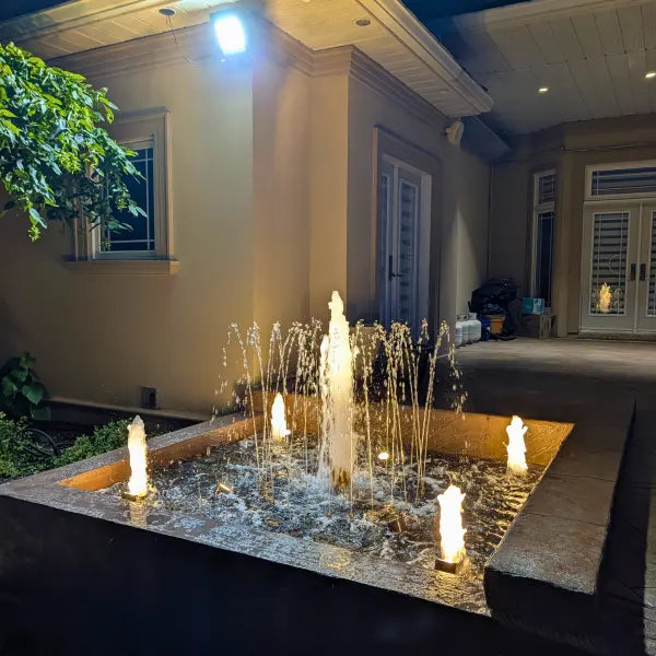 An outdoor rectangular fountain pool at night with multiple illuminated candle-style jets and water arcs, set in front of a house entryway.