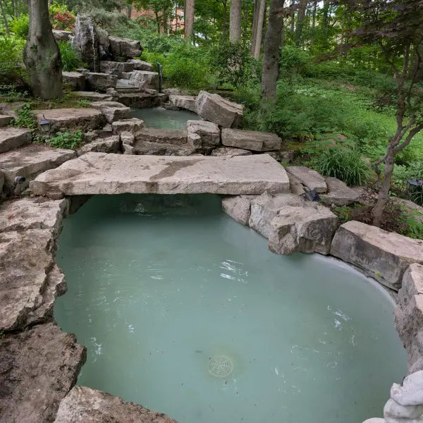 A natural stone hot spring or geothermal pool with pale greenish water, surrounded by large rocks and a stone bridge, set in a lush green forest environment.
