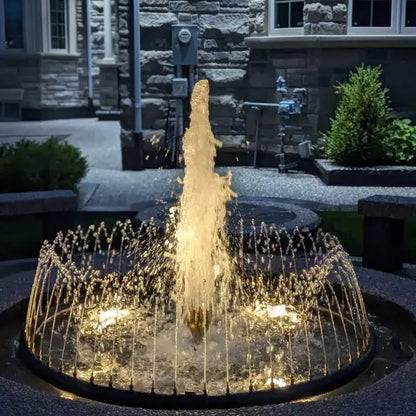 A circular outdoor fountain with a tall central jet of water illuminated in warm yellow light, surrounded by smaller spraying arcs. The fountain is set in a stone courtyard with landscaping and building architecture visible in the background.