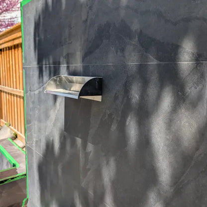 A stainless steel outdoor wall-mounted water spillway installed on a dark gray stone tile wall, with natural tree shadows cast across the surface.