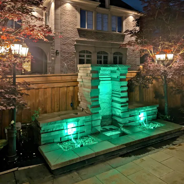 A modern stone waterfall fountain with integrated green LED lighting, set in a backyard patio area at night with a brick house and trees in the background.