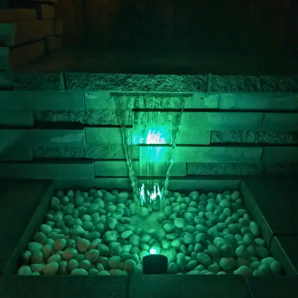 A small outdoor waterfall feature with water flowing into a pebble basin, illuminated by green lighting at night.